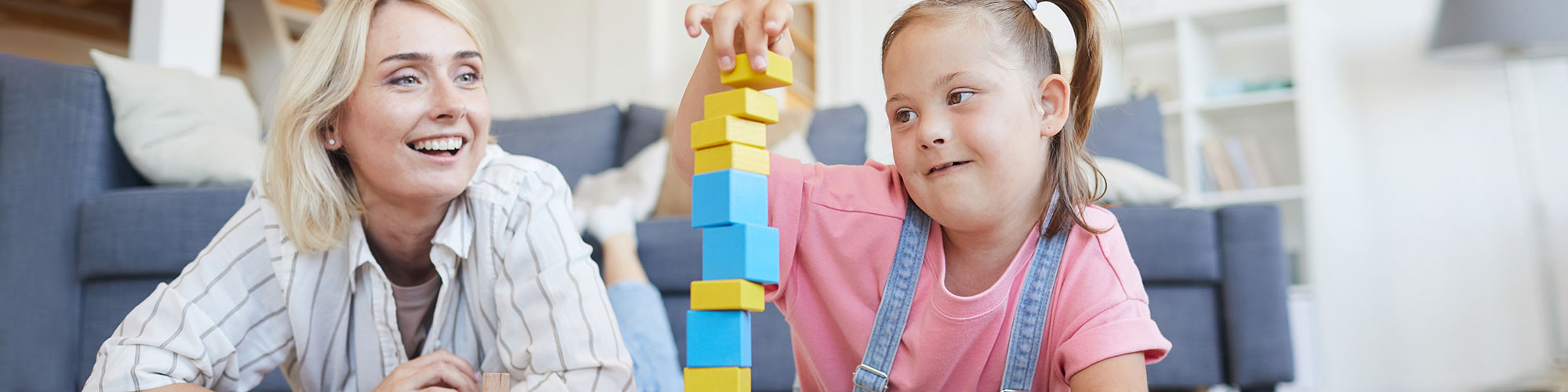 Eine Frau und Mädchen mit Down-Syndrom spielen gemeinsam am Boden und bauen einen Turm aus bunten Klötzchen.