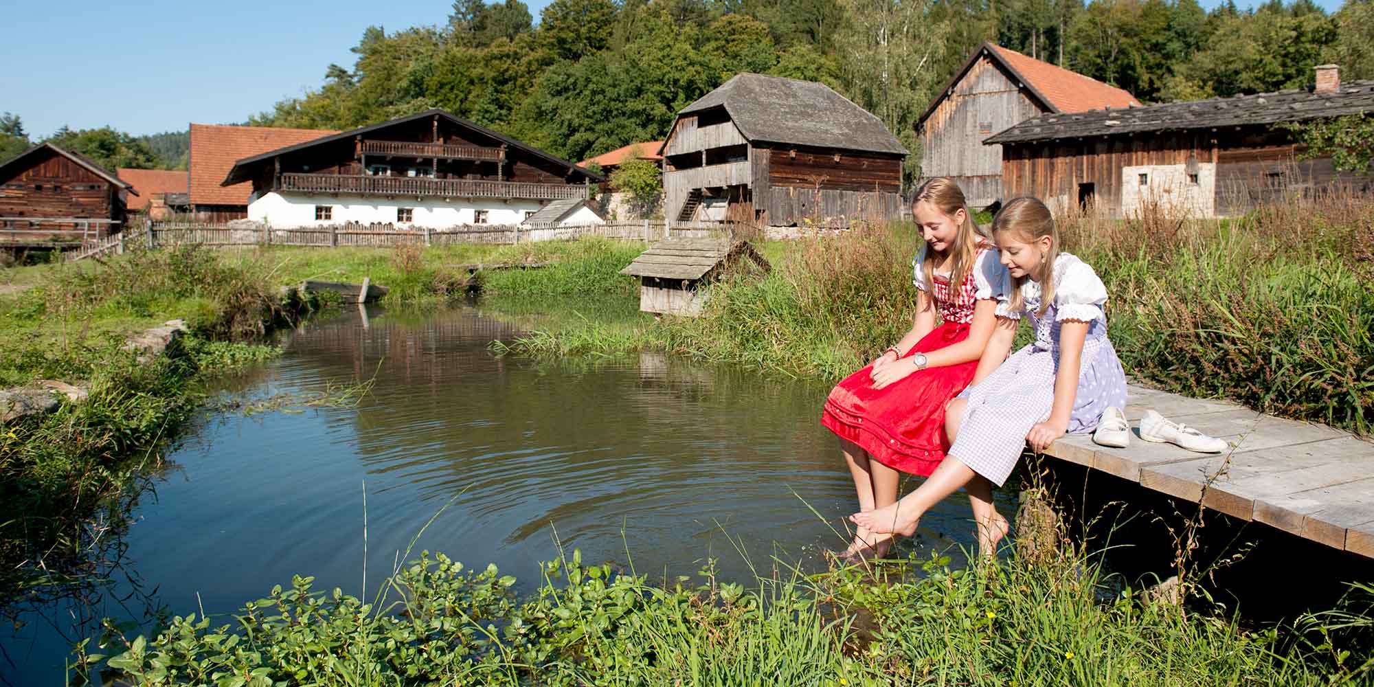 Zwei Mädchen in Tracht sitzend am Teich im Museumsdorf Bayrischer Wald